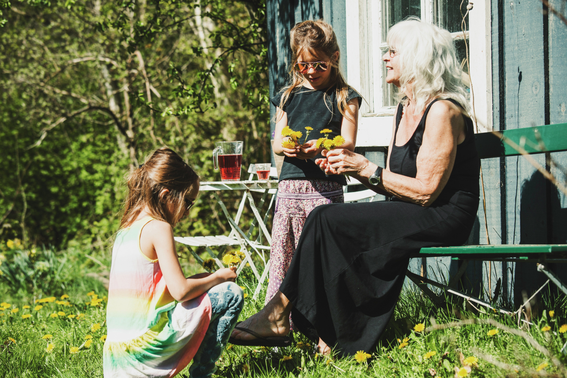 Bedstemor med børnebørn sidder uden for i solen ved sommerhus, fotografi