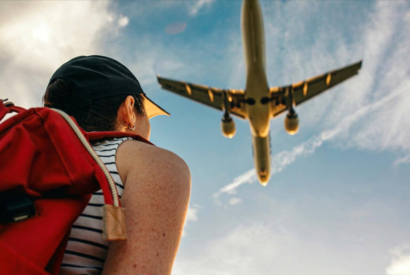 En person med rygsæk står udendørs og ser et lavtflyvende fly passere hen over himlen.